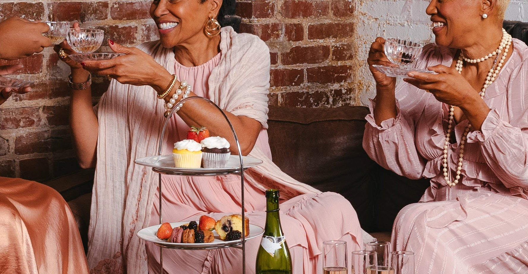 Group of black women sitting together in a cozy room with a brick wall and wooden floor with an inviting tea party setting.